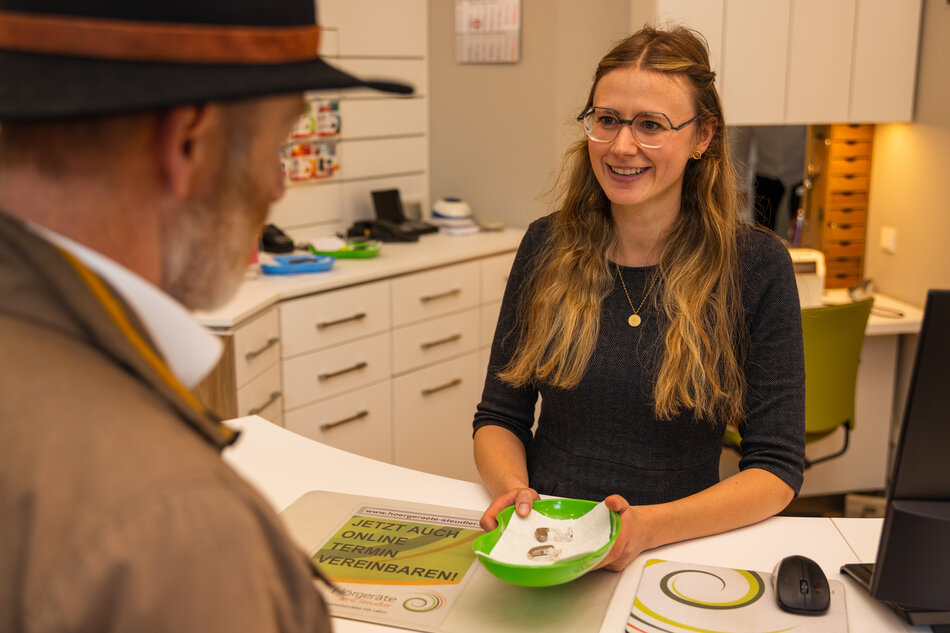 Eine Mitarbeiterin von Hörgeräte Jens Steudler zeigt einem Kunden am Tresen eine Schale mit Hörgeräten